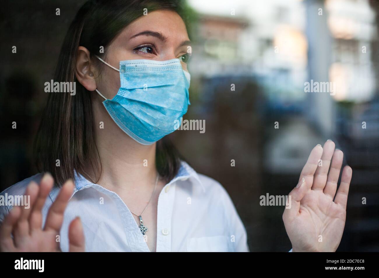 Schöne junge Frau trägt Gesichtsmaske, Blick durch Fenster draußen, Hände gegen Glas, Coronavirus COVID-19 globale Pandemiekrise, zu Hause bleiben Stockfoto