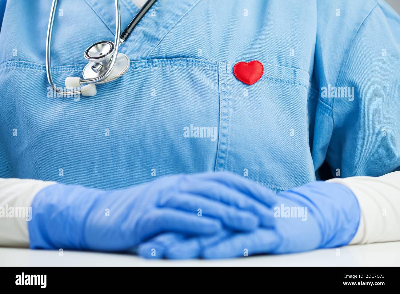 Arzt in blauer Uniform trägt ein Stethoskop und kleine rote Herz Abzeichen Pin, Detail Nahaufnahme, Hausarzt oder Kardiologe im Büro, sitzt am Schreibtisch Stockfoto
