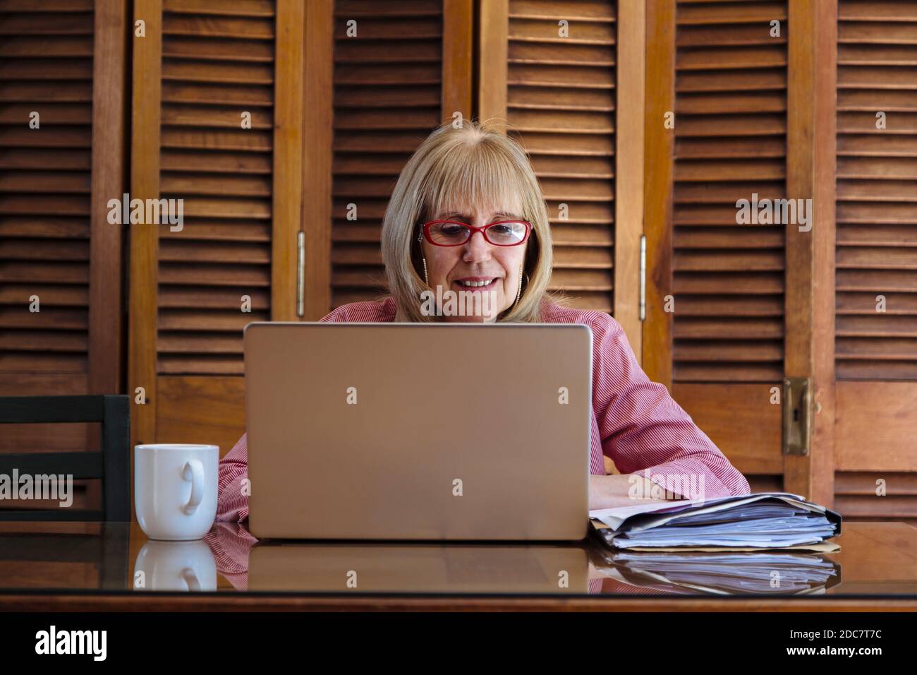 Ältere Frau, die mit einem Laptop arbeitet und Papiere sucht. Sie ist drinnen in einem Holztisch mit einem silbernen Computer. Vorderansicht. Stockfoto