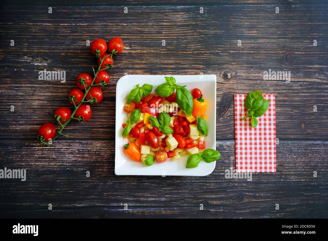 Frische Zutaten für einen gesunden Salat auf einem weißen quadratischen Teller: Tomaten, Käsewürfel, Paprika und Basilikum. Dunkler Holzhintergrund mit Serviette. Stockfoto