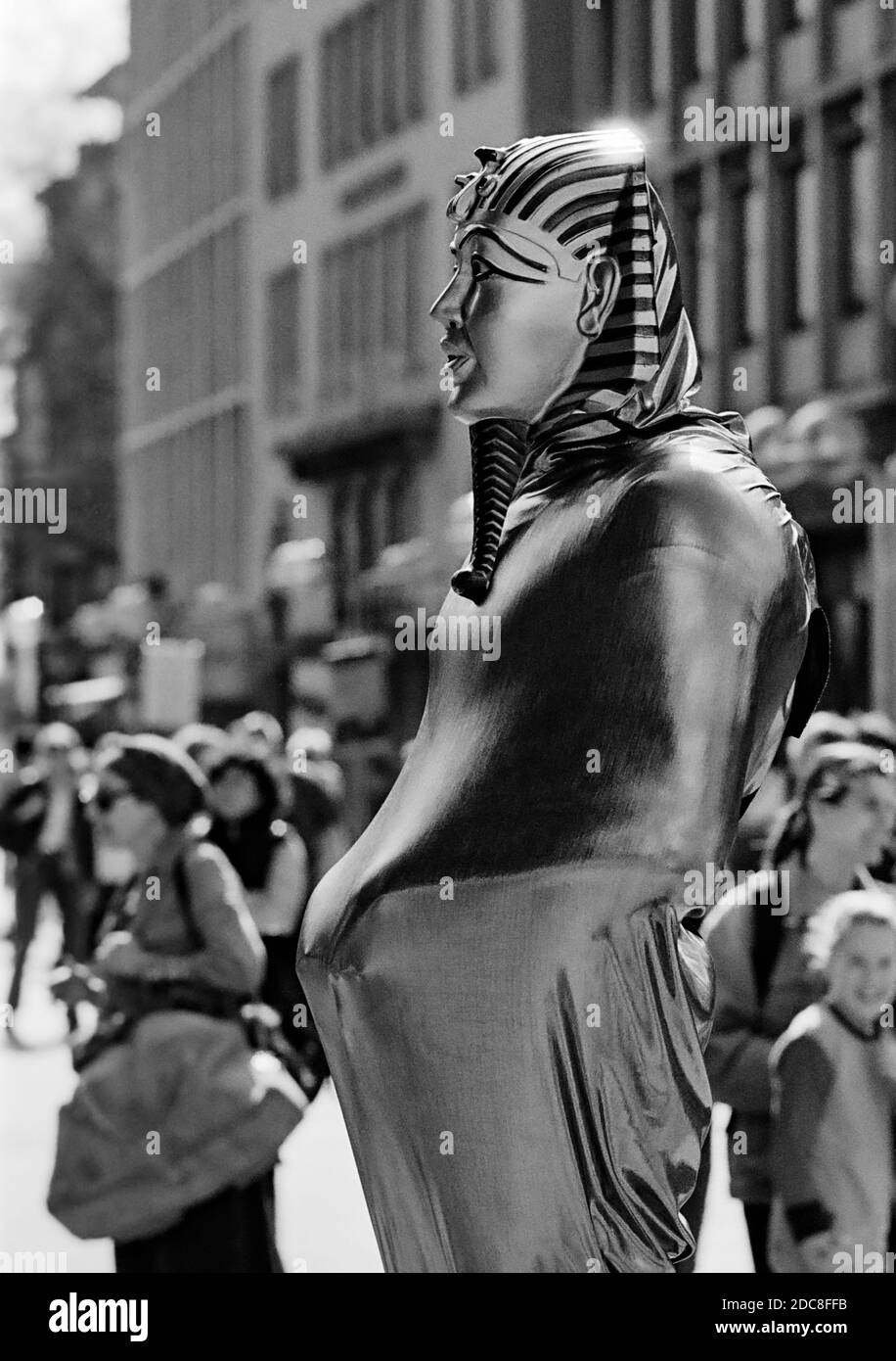 Showman in der Kölner Fußgängerzone in der Nähe der Kuppel trägt eine Maske aus Tutanchamun und ein eng anliegender silberner Kostüm, der in der Sonne scheint. Stockfoto