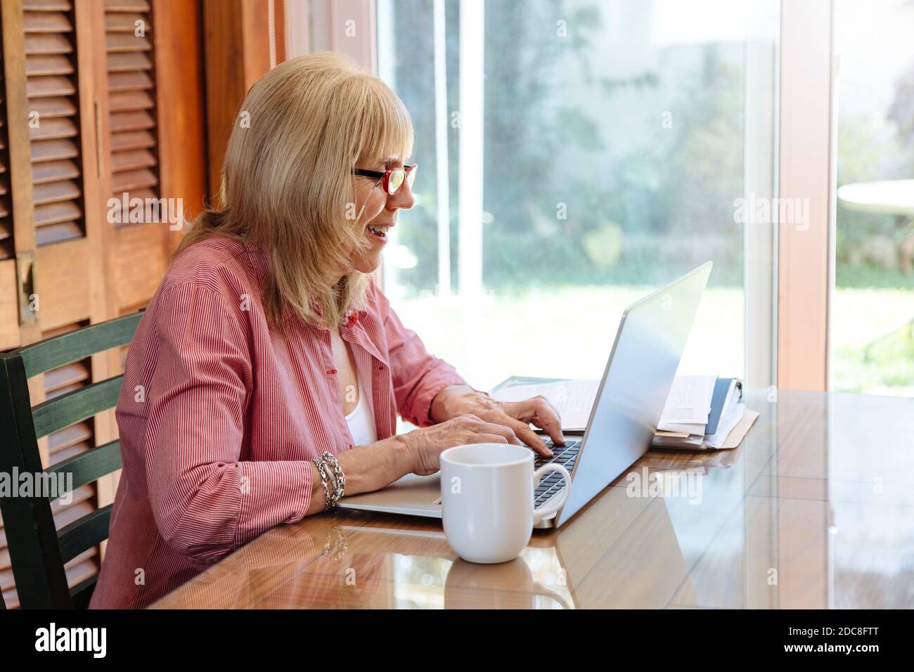Lustige ältere Frau mit Laptop. Lächelnde Großmutter auf einem Holztisch mit einem silbernen Computer. Kaffeetasse beiseite. Stockfoto