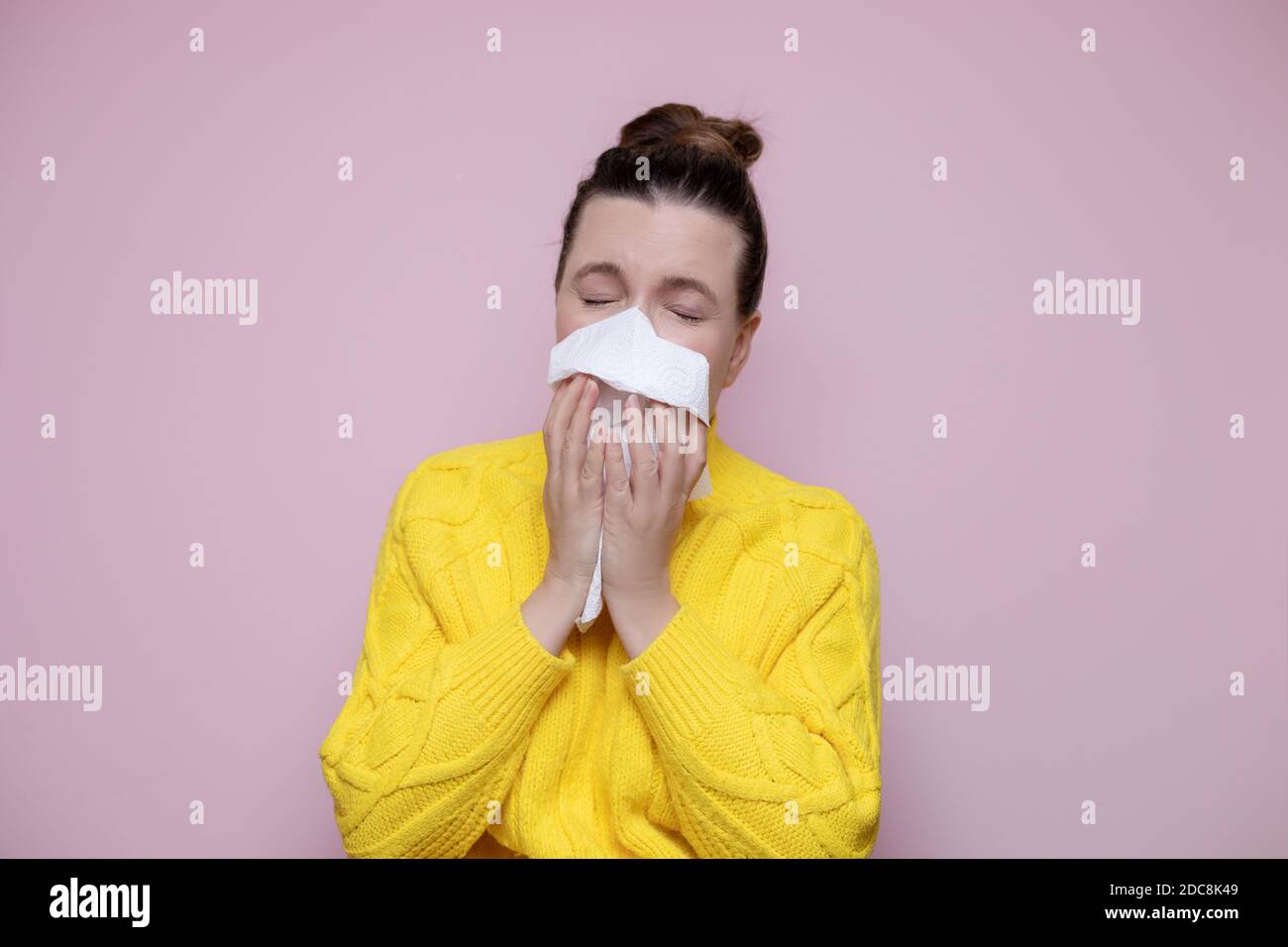 Foto kranke Frau in einem Pullover niest in eine Serviette auf einem rosa Hintergrund, allergische Rhinitis. Das Konzept von Erkältungen, saisonalen Erkrankungen, Grippe. Stockfoto