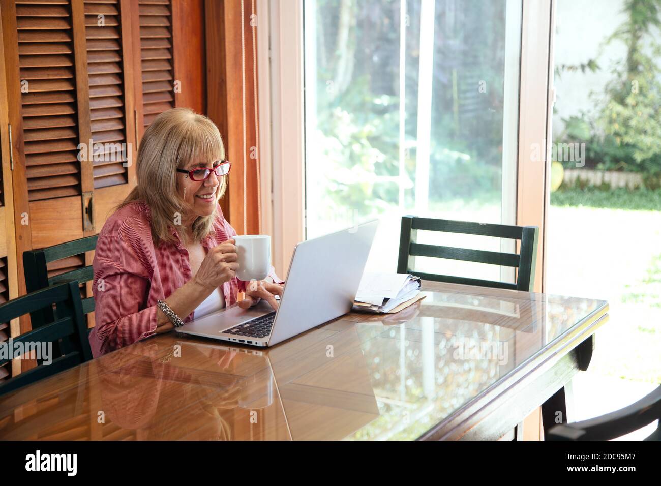 Ältere Frau mit Laptop und Kaffeetasse in der Hand. Lächelnde Großmutter auf einem Holztisch mit einem silbernen Computer. Stockfoto