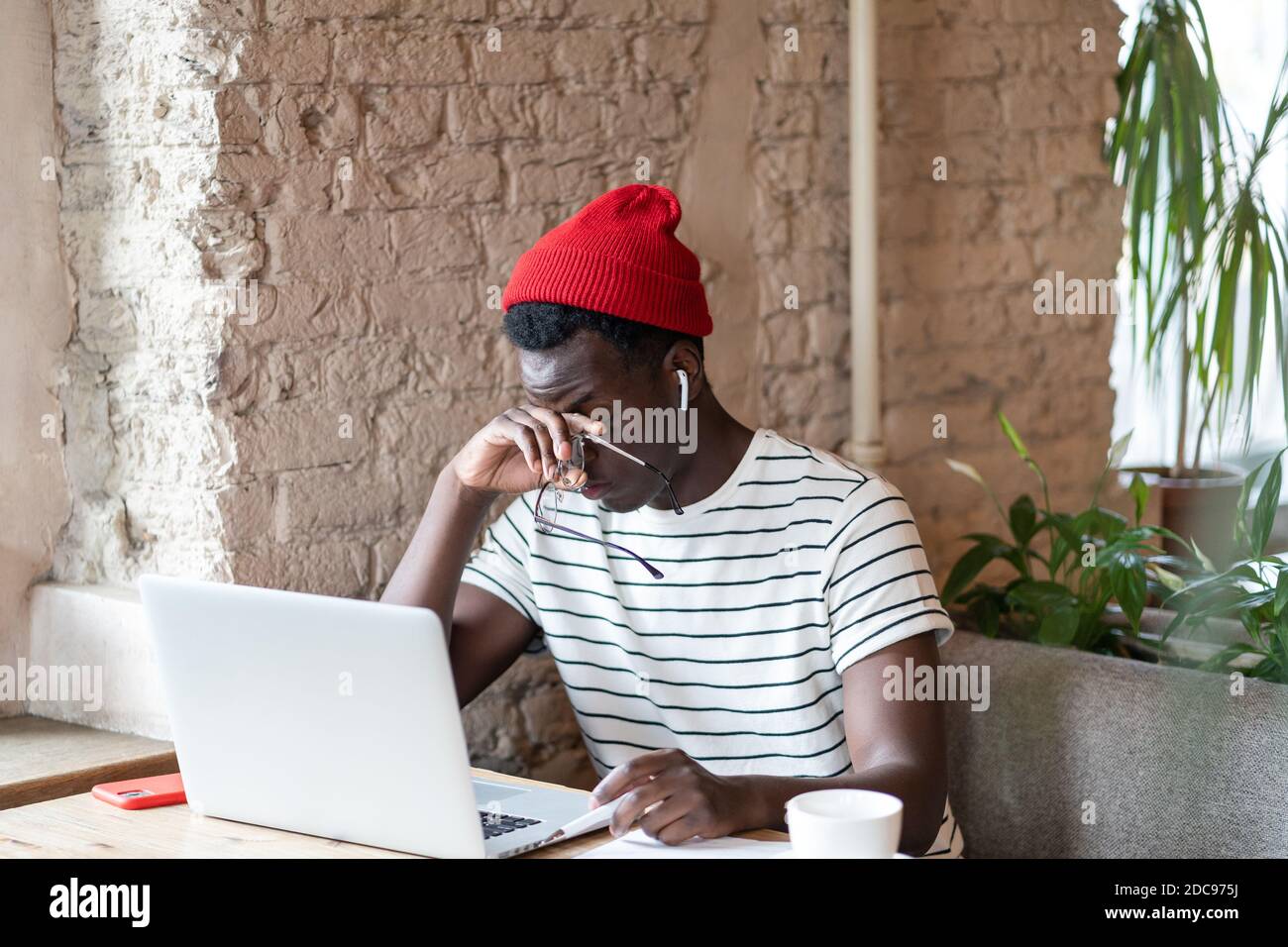 Millennial Afro-American Mann in roten Hut tragen drahtlose Kopfhörer, hält Brille, reiben seine Augen, fühlt sich müde nach der Arbeit am Laptop im Café, taki Stockfoto