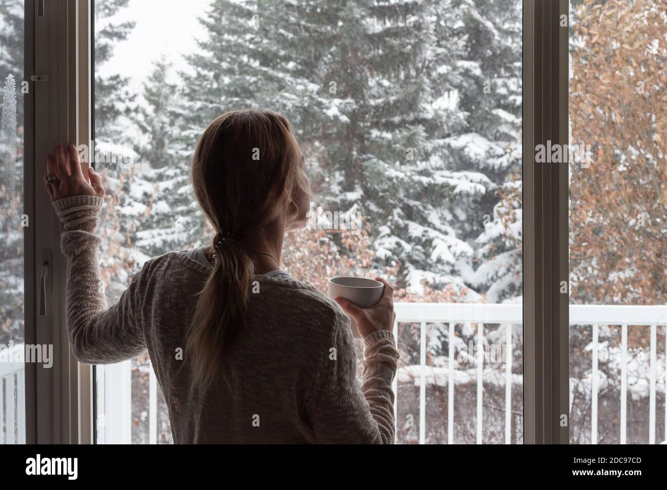 Frau mit Blick aus dem Fenster auf den Winter, schneebedeckte Bäume, mit Kaffeebecher in der Hand Stockfoto