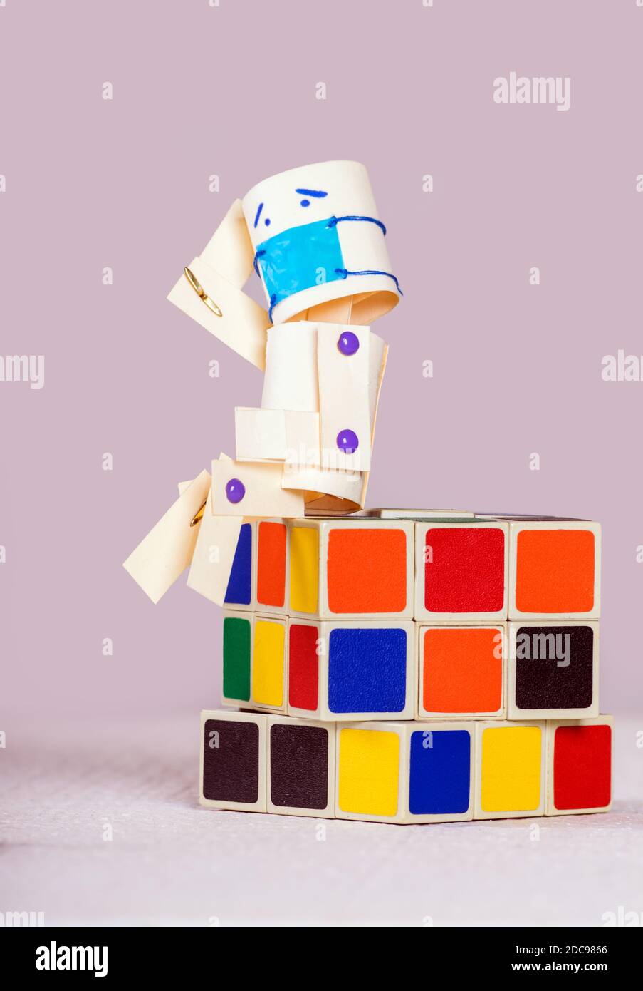 Papierpuppe mit einem müden Ausdruck trägt eine chirurgische Maske sitzt auf einem Standard Rubik Würfel. Stockfoto