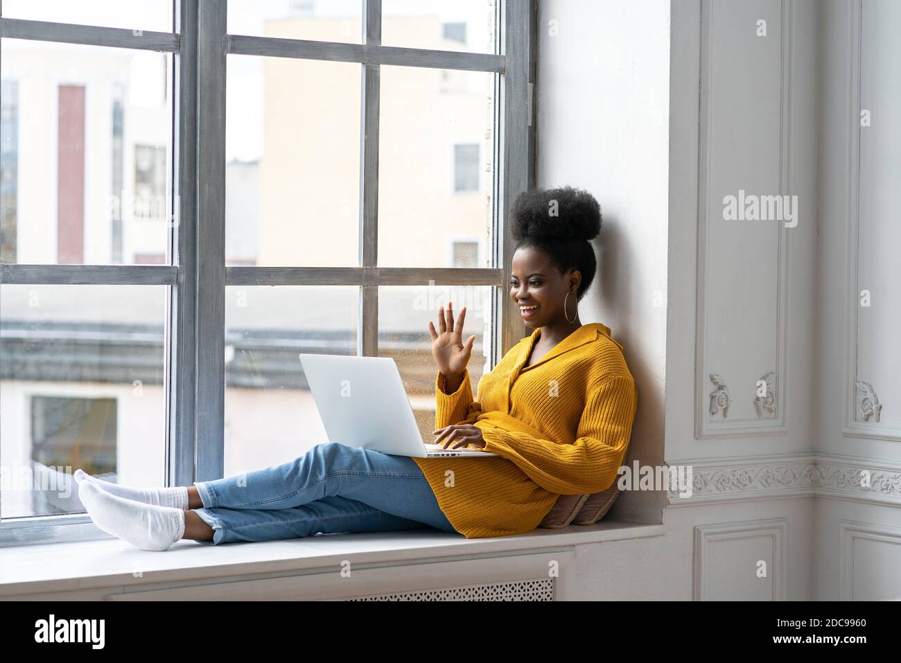 Lächelnde afroamerikanische Millennial Frau mit afro Frisur in gelber Strickjacke sitzt auf Fensterbank, ruht, Blick auf Kamera Webcam, sagen hallo, Stockfoto