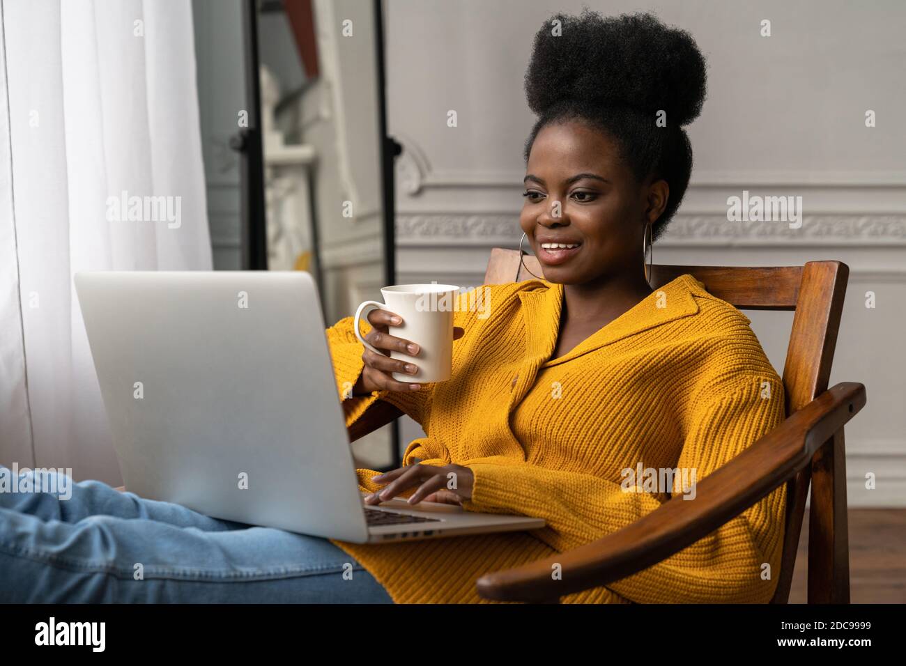 Happy Afro-amerikanische Millennial Frau mit Afro Frisur tragen gelbe Strickjacke ruhen, sitzen auf einem Stuhl, beobachten Webinar, arbeiten online auf Laptop, t Stockfoto