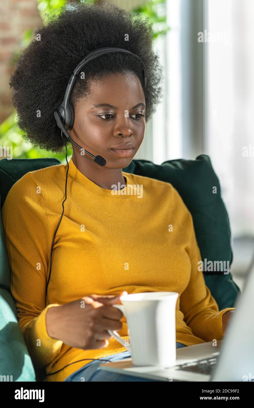 Afro-amerikanische Beraterin Frau tragen Kopfhörer Fernarbeit am Computer während Lockdown, trinken Tee oder Kaffee, halten Tasse. E-Business, Kundenbetreuung Stockfoto