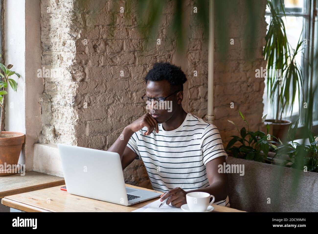 Fokussierte Millennial Black man Student in T-Shirt Stripped Brille tragen, beobachten pädagogische Webinar auf Laptop, Remote-Online-Arbeit im Café, Studium, Stockfoto