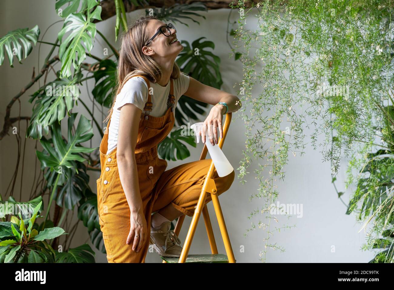 Glückliche Gärtnerin Frau hält Pulverisierspray, lächelt, macht eine Pause von der Arbeit, sitzt auf der Trittleiter, trägt Brille und orange Overalls. Grün Stockfoto