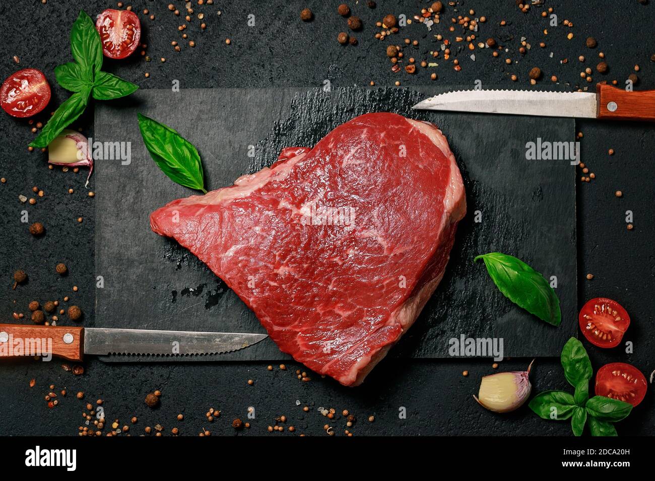 Rohes Rindersteak auf einem Steinschneidebrett. Gras gefüttertes Fleisch mit Gewürzen und Kräutern auf dunklem Hintergrund. Draufsicht Stockfoto