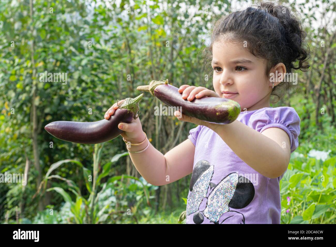 Portrait eines kleinen Mädchens, das mit zwei frischen Auberginen spielt, die aus dem Garten gepflückt wurden, gesundes Lifestyle-Konzept Stockfoto