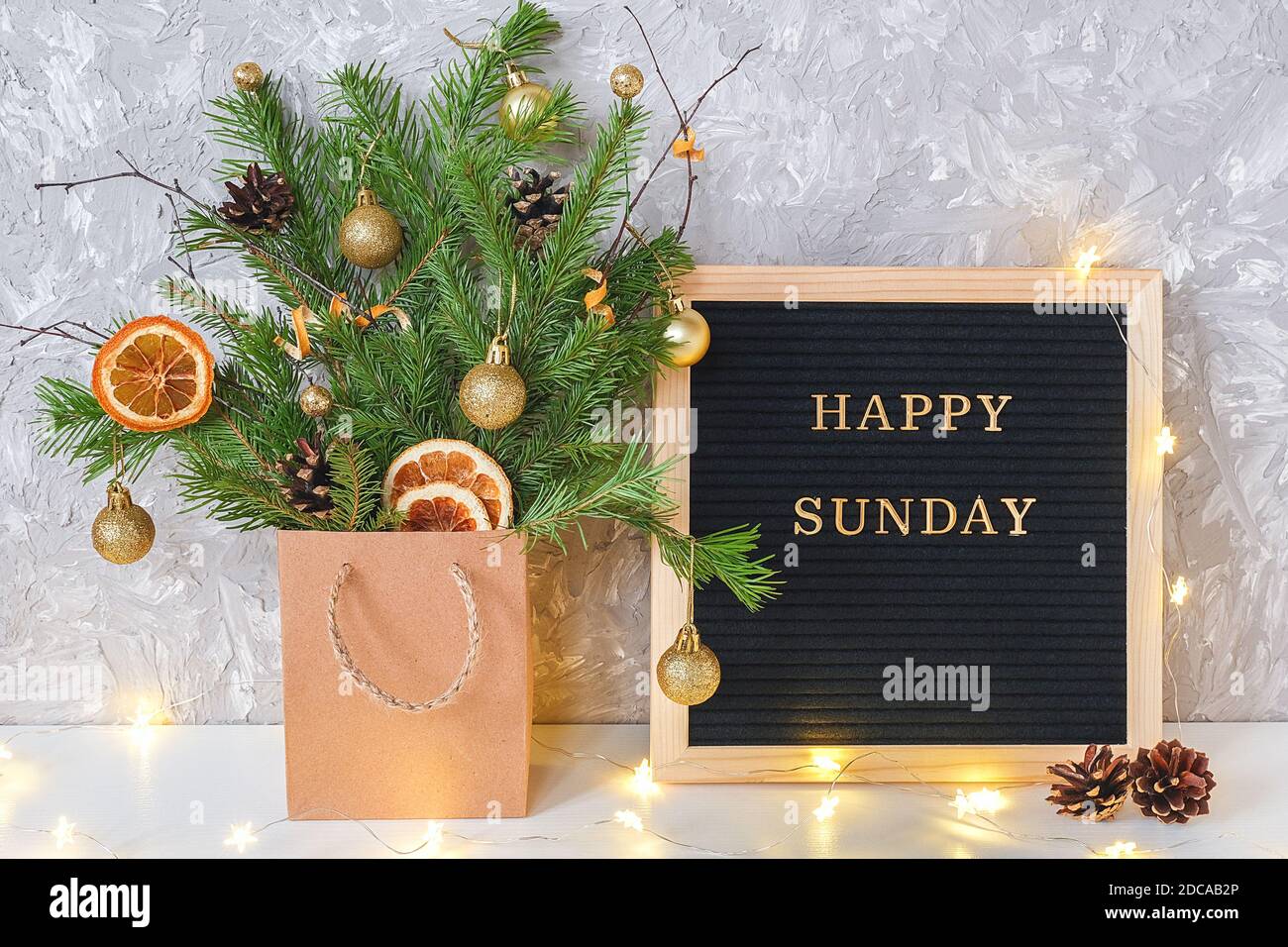 Happy Sunday Text auf schwarzem Briefbrett und festlicher Blumenstrauß aus Tannenzweigen mit weihnachtsdekor in Bastelpackung und Lichtlampe Girlande auf Tisch. Tem Stockfoto