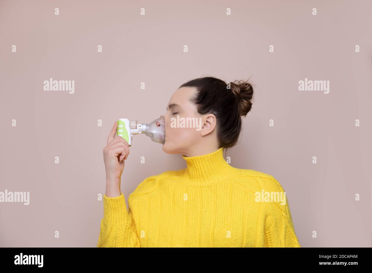 Eine 40-jährige Frau in einem gelben Pullover auf rosa Hintergrund inhaliert die oberen Atemwege, die Lunge mit Hilfe eines kabellosen Verneblers. Augen Stockfoto