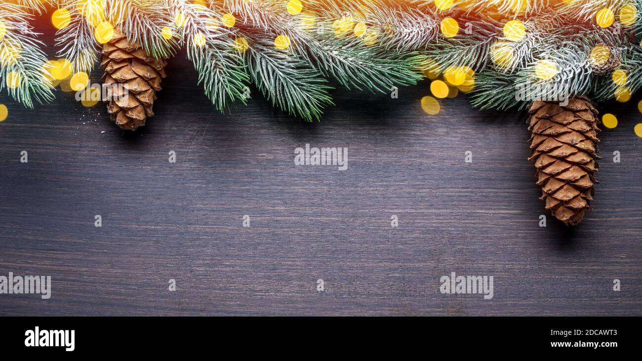 Schneebedeckter Tannenzweig mit Tannenzapfen und verschwommenen Weihnachtslichtern auf Holztisch. Weihnachten oder Neujahr Urlaub Hintergrund. Draufsicht. Stockfoto