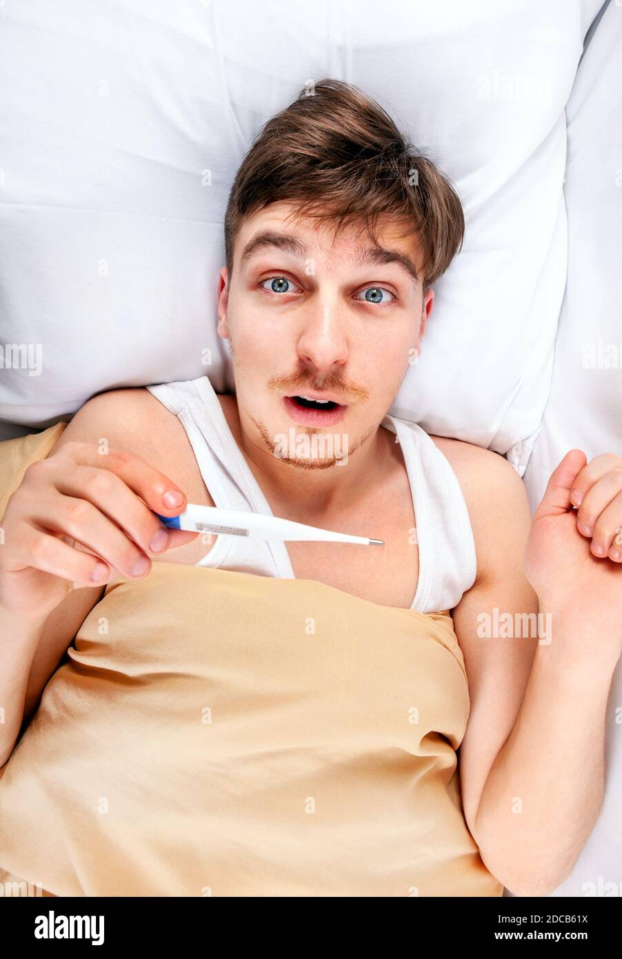 Schockierter junger Mann mit einem Thermometer im Bett bei Das Zuhause Stockfoto