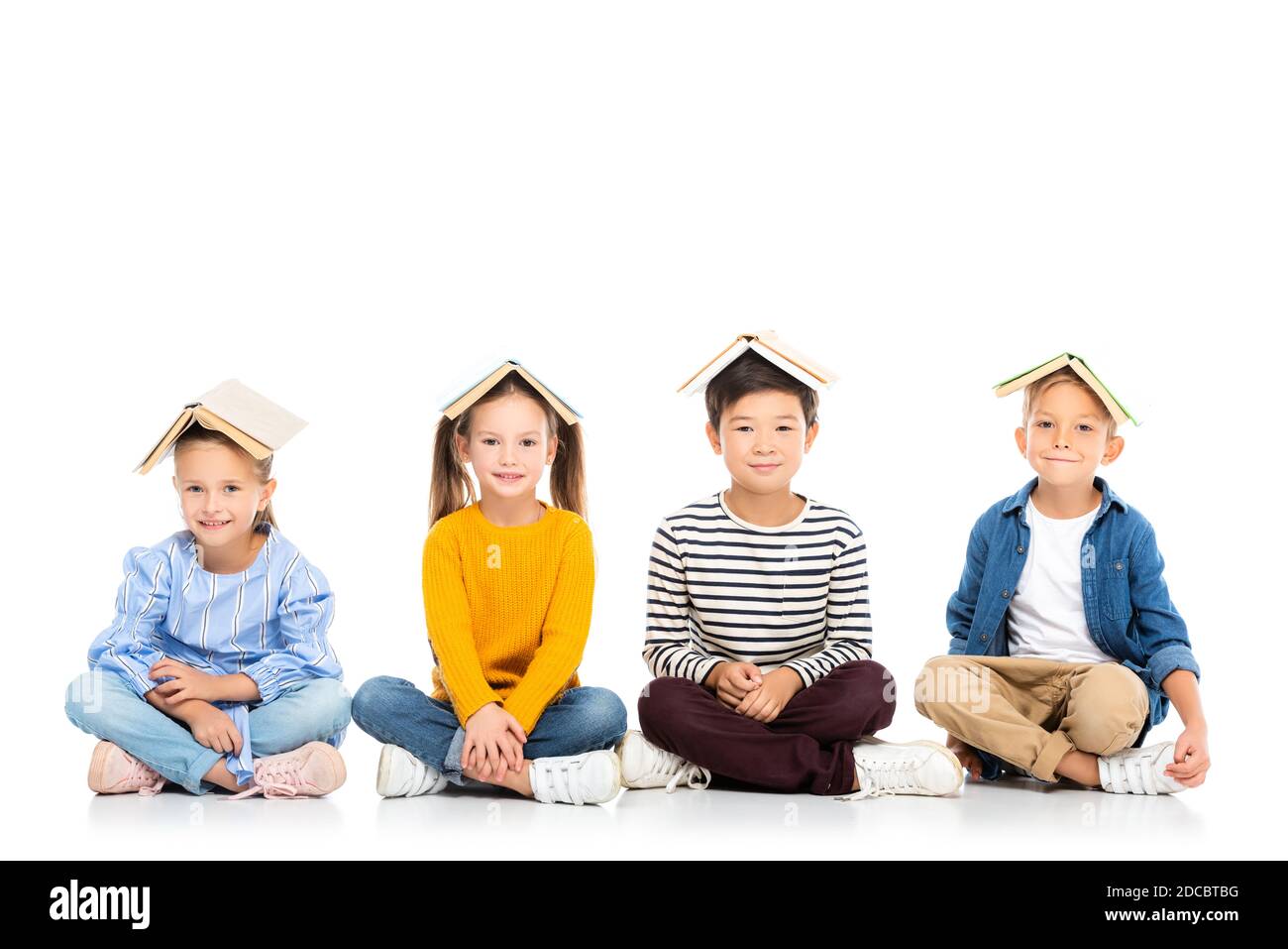 Lächelnde multiethnische Kinder mit Büchern auf Köpfen sitzen auf weiß Hintergrund Stockfoto