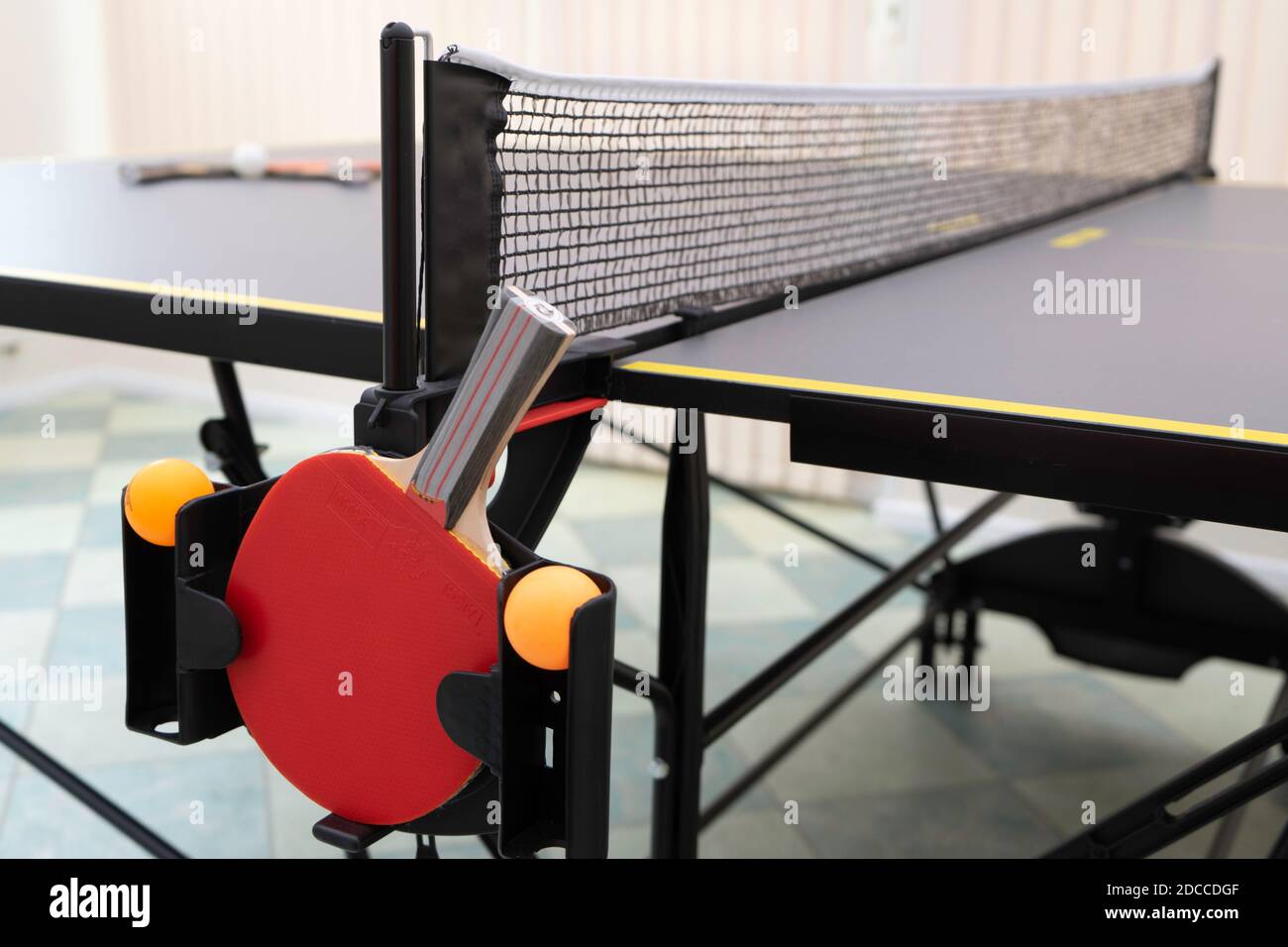 Eine schräge Seitenansicht einer Tischtennisplatte und Netz mit einem roten Tischtennisschläger und zwei orangefarbenen Tischtennisbälle in einem Halter und zwei weitere Tische tenni Stockfoto