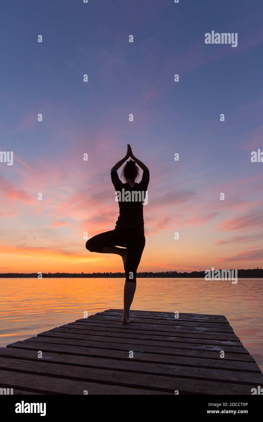 Frau üben stehende Haltung Vrikshasana / Baum Pose, ein Bein ausbalancieren Asana des mittelalterlichen Hatha Yoga auf Steg am See bei Sonnenuntergang Stockfoto