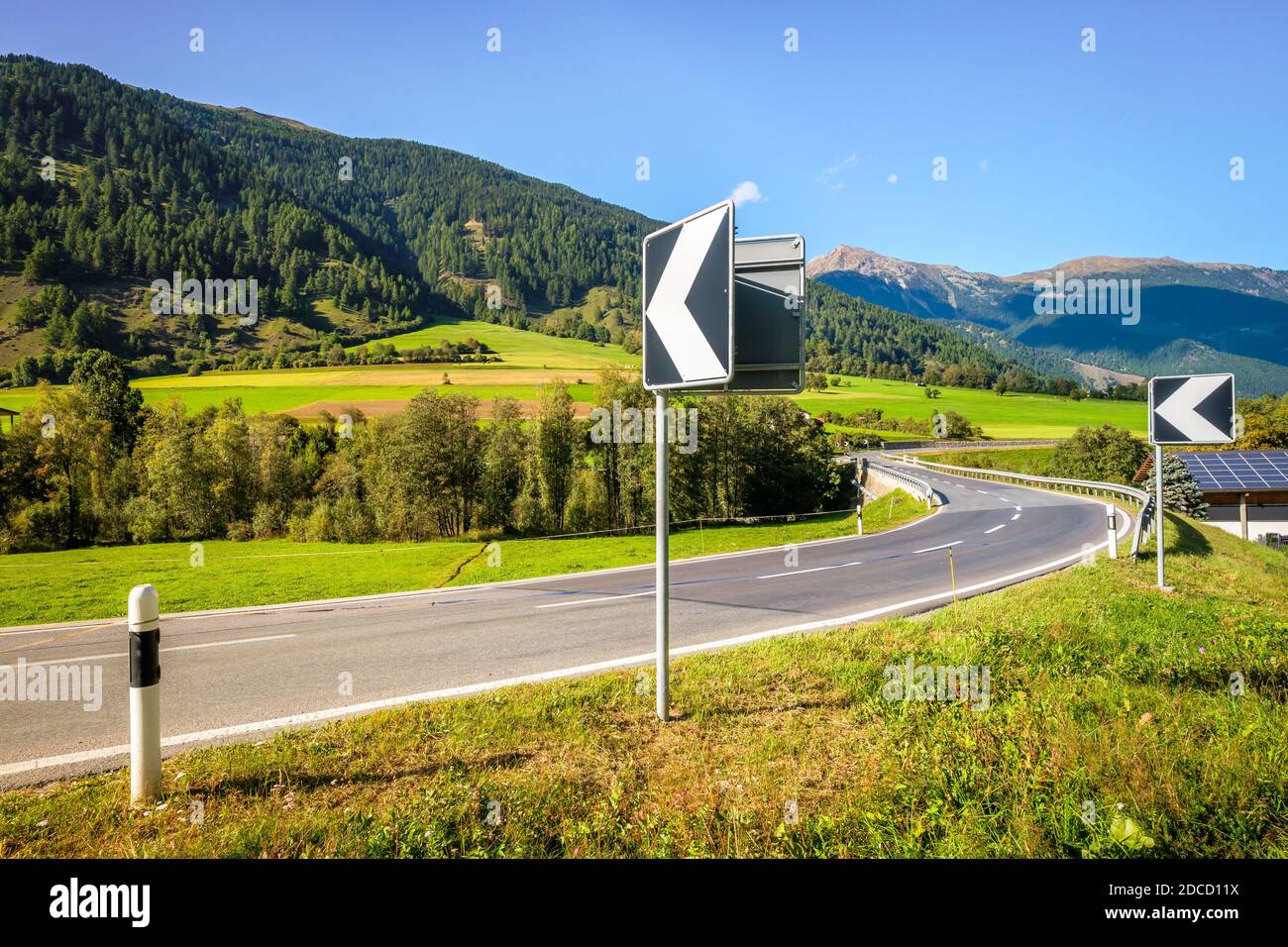 Straßenschilder weisen auf eine Biegung an einer Brücke über den Fluss Rom im Schweizer Val Müstair hin. Die Straße führt zum Dorf Müstair und dann zur italienischen Grenze. Stockfoto