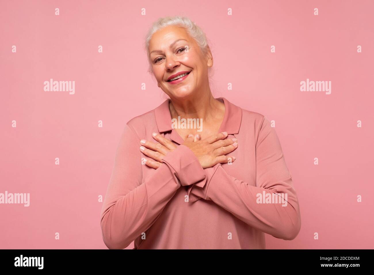 Ältere Frau bekommt immer wieder Komplimente. Entzückte glücklich charmante alte Dame mit weißen Haaren lächelt an der Kamera. Studio auf rosa Wand aufgenommen. Stockfoto