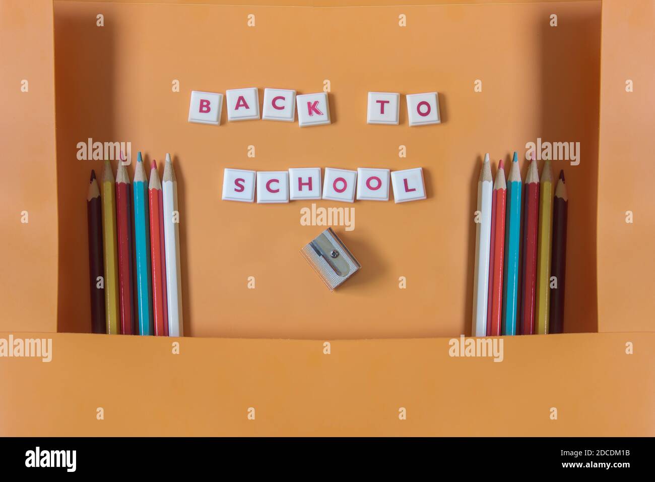 Eine offene orangefarbene Mappe mit Farbstiften an den Seiten, einem Bleistiftspitzer und Mosaikbuchstaben, die die Phrase 'Back to School' bilden. Stockfoto