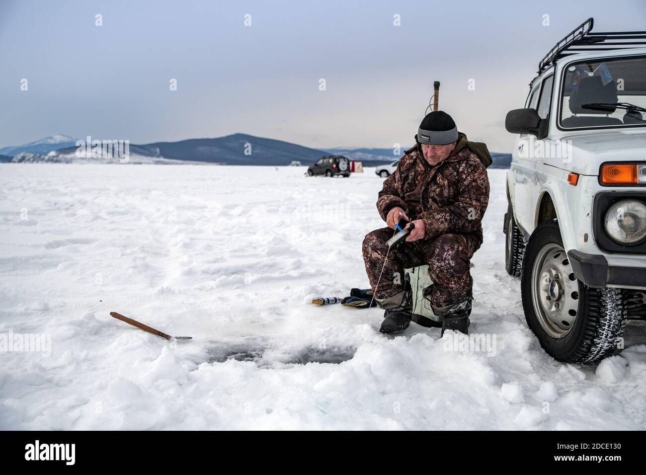 Baikalsee, Sibirien, Russland - 05. März 2020: Wintereisfischen. Ein Fischer fischt auf Spinten in der Nähe von Zelten und Autos auf dem Baikalsee auf einem Marsch d Stockfoto
