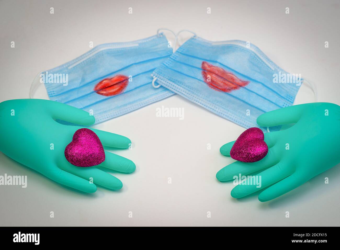 Es gibt zwei medizinische Masken mit roten Lippen auf sie gemalt. Im Vordergrund sind grüne medizinische Handschuhe, die jeweils ein scharlachrot Herz tragen. Selektiver Fokus Stockfoto