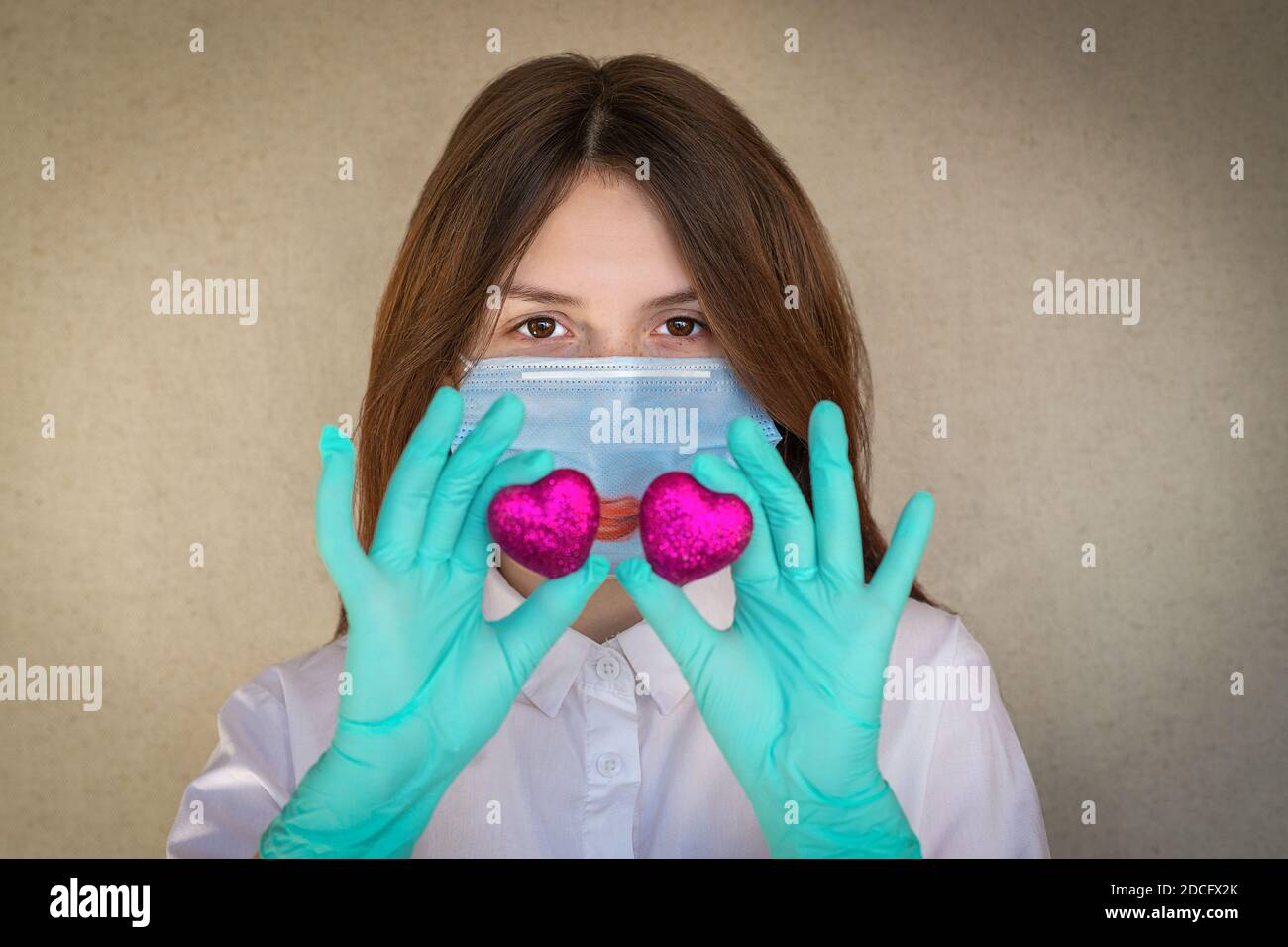 Ein dunkelhaariges Mädchen von 14-15 Jahren, das eine medizinische Maske und Handschuhe trägt, hält zwei scharlachrote Herzen vor sich. Selektiver Fokus Stockfoto