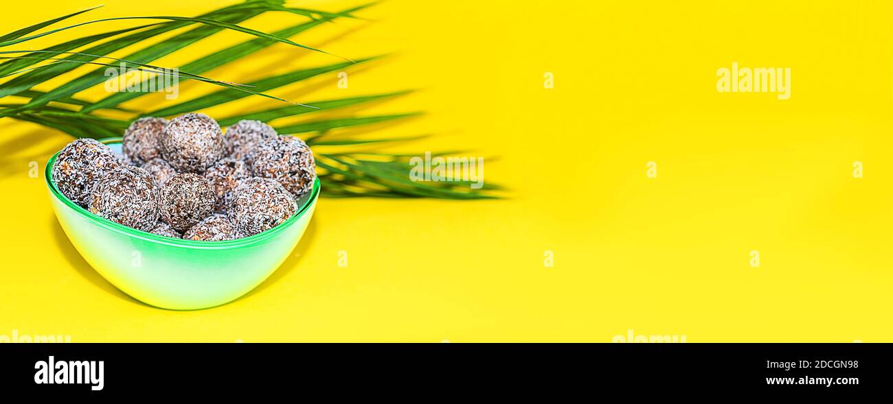 Protein Erdnussbutter Energie Bisse oder Energiebälle. Hausgemachte rohe Schokoladentrüffel mit Nüssen und in Kokosflocken auf Teller mit Palmblatt auf gelbem Hintergrund. Stockfoto