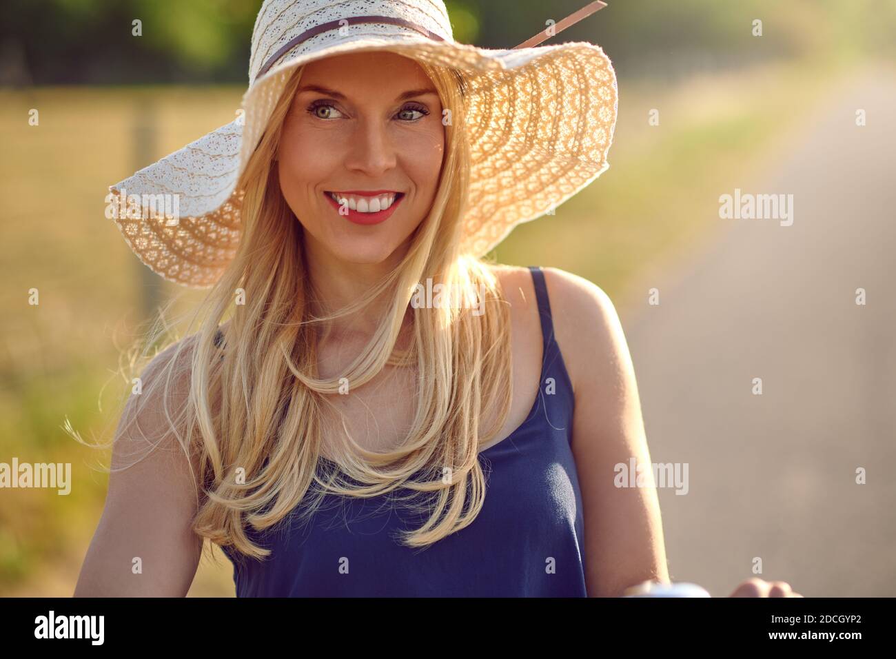Attraktive Frau mittleren Alters in einem breiten Strohsonnenhut Und blau Sommer Top lächelt an der Kamera, wie sie Blicke beiseite auf eine sonnige Landstraße Stockfoto