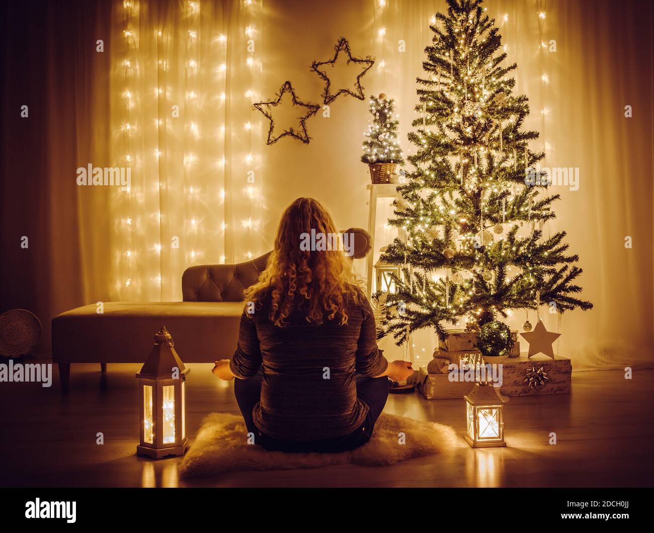Frau sitzen auf Schaffell Teppich und meditieren. Ruhiges, entspannendes Weihnachtskonzept. Geschmückter Weihnachtsbaum mit Eiszapfen und Schneeflocken-Ornamenten. Stockfoto