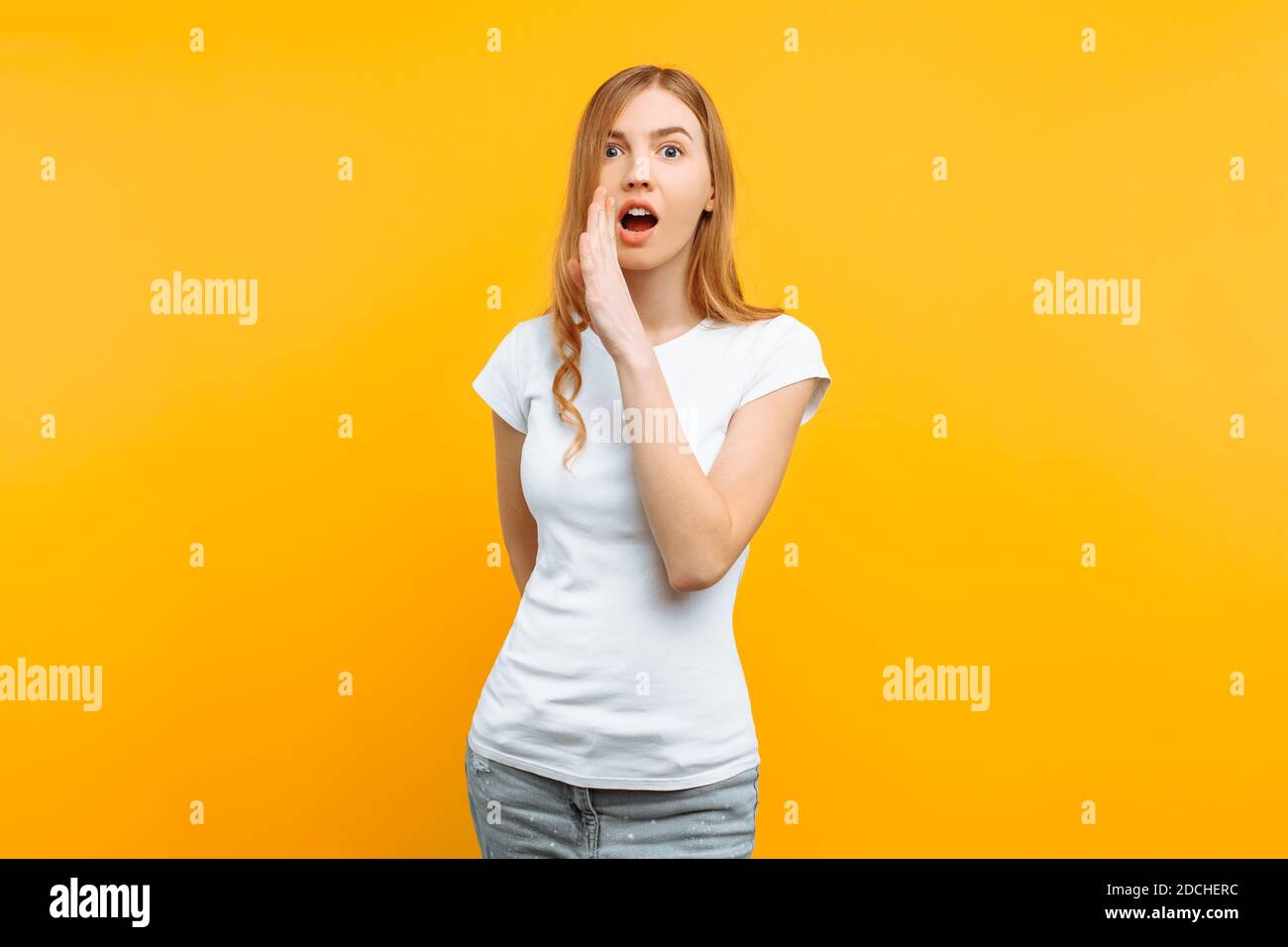 Junge schockierte Frau in weißem T-Shirt, hält eine Hand in der Nähe des Mundes und erzählt ein Geheimnis, auf einem gelben Hintergrund Stockfoto