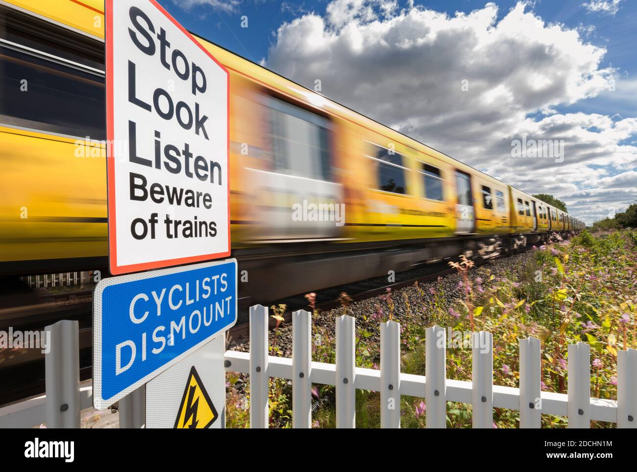 Merseyrail Zug vorbei an einem öffentlichen Fußgängerübergang mit Geschwindigkeit Bewegungsunschärfe mit dem Stop-Look-Listen-Warnschild und Radfahrer demontieren Schild Stockfoto