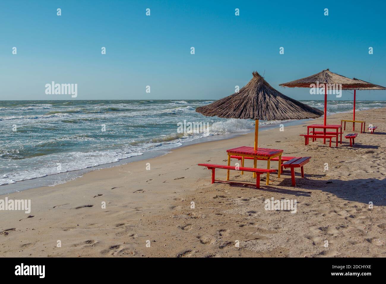 Sommerlandschaft: Tisch, Bänke und Sonnenschirm am Strand. Goldener Sand, stürmisches und winkendes Meer und blauer Himmel. Stockfoto