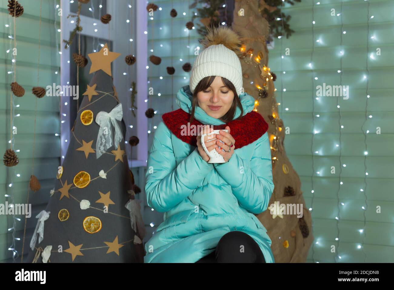 Junge Frau in warmen Hut, Schal und Daunenjacke trinkt Tee auf dem Hintergrund der Weihnachtsdekoration Stockfoto