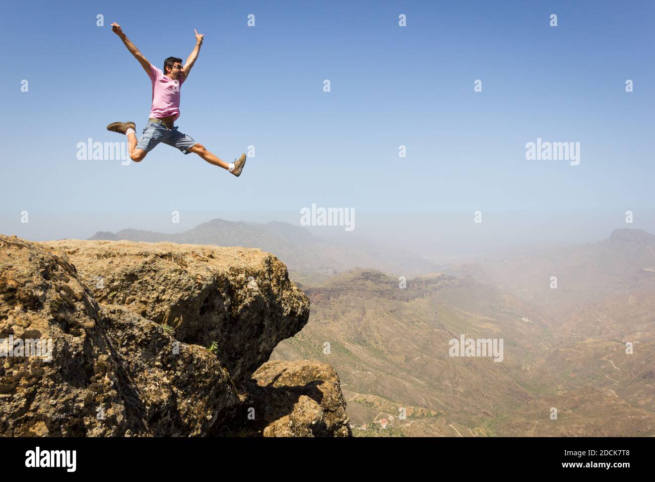 Tapferer Mann auf pinkem T-Shirt, der hoch oben am Felsrand im Naturpark Roque Nublo auf Gran Canaria springt. Junger Kletterer feiert auf dem Berggipfel Stockfoto