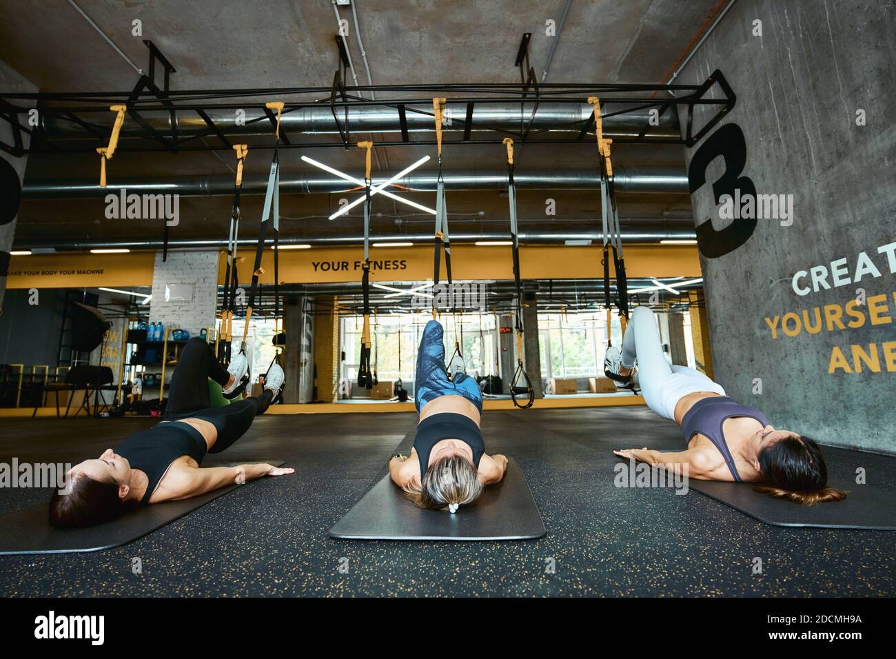 Drei junge kaukasische Sportlerinnen in Sportbekleidung liegen auf dem Boden und trainieren mit Fitness trx Gurten im Industrial oder Crossfit Gym. Sport, Menschen arbeiten, Wellness und gesunde Lebensweise Stockfoto