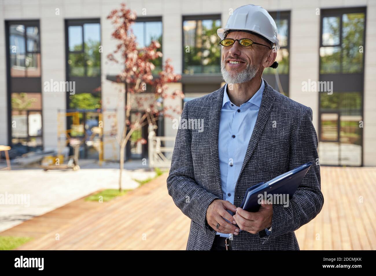 Städtisches Design. Erfolgreicher Architekt mittleren Alters, Ingenieur Manager trägt Helm lächelnd, im Freien stehend und hält eine Blaupause in seinem Stockfoto