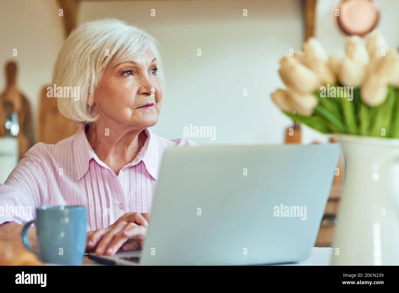 Die Taille nach oben lächelnde ältere Frau sitzt am Tisch mit Laptop am Morgen, während sie weg schaut. Inländisches Lifestyle-Konzept Stockfoto
