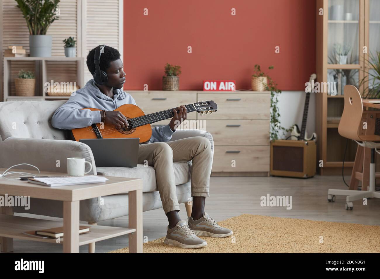 Ganzkörperportrait des jungen afroamerikanischen Mannes, der akustische Gitarre spielt, während er auf der Couch sitzt und zu Hause Musik macht, Raum kopieren Stockfoto