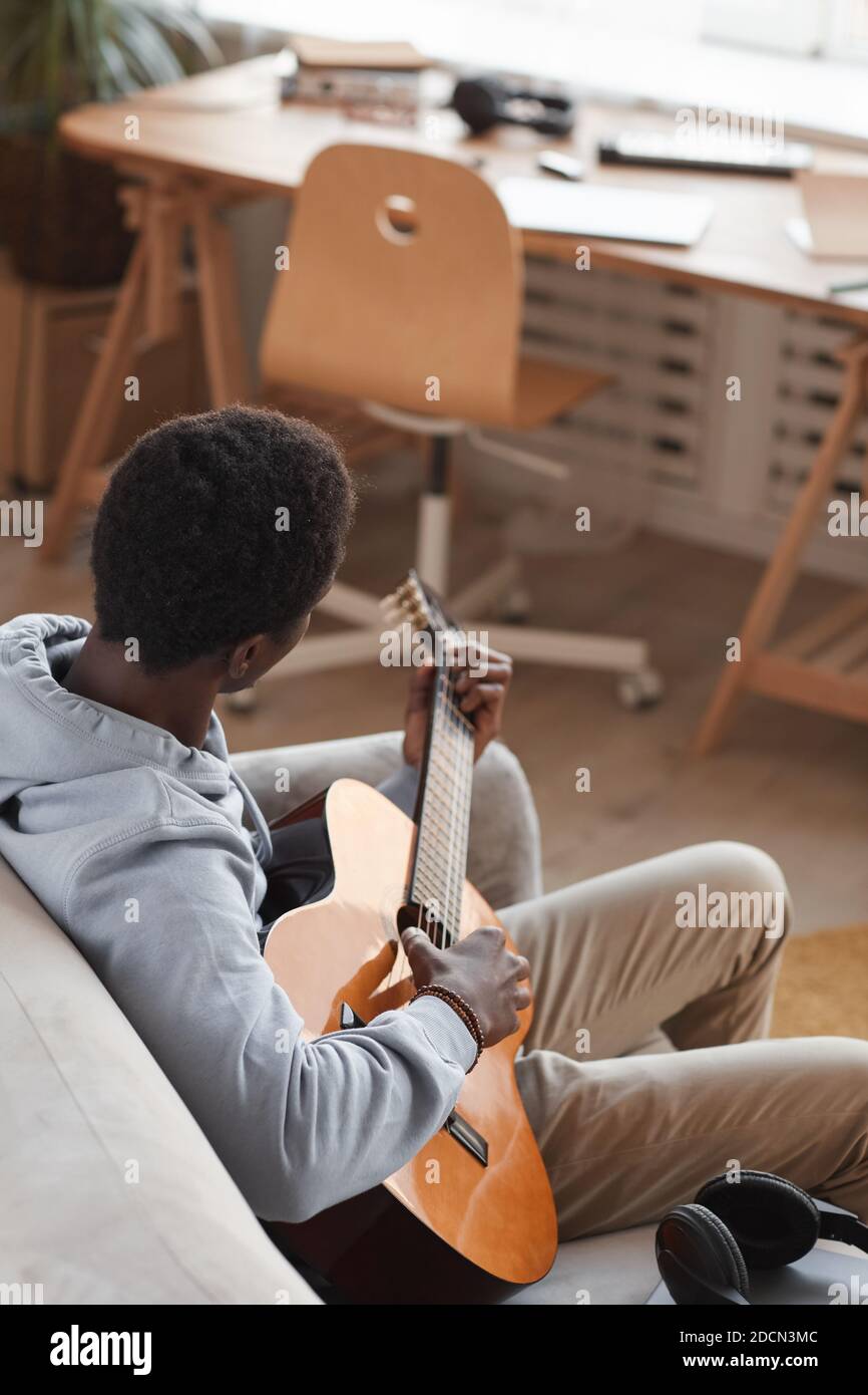 Vertikales Hochwinkelporträt des jungen afroamerikanischen Mannes, der akustisch spielt Gitarre und beim Sitzen auf der Couch zu Hause Stockfoto
