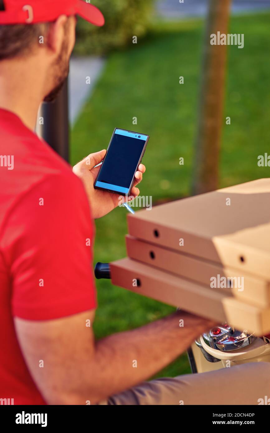 Rückansicht eines Liefermanns in roter Uniform, der auf einem Roller sitzt und mit dem Smartphone unterwegs ist, während er Pizza liefert. Konzept für die Lebensmittelzustellung Stockfoto
