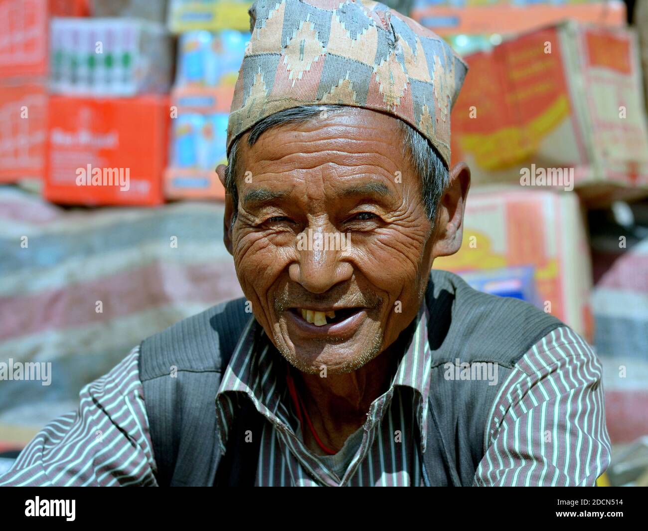 Der ältere nepalesische Tamang-Händler trägt einen traditionellen nepalesischen Hut (dhaka topi) und lächelt auf dem Wochenmarkt vor seiner Ware für die Kamera. Stockfoto