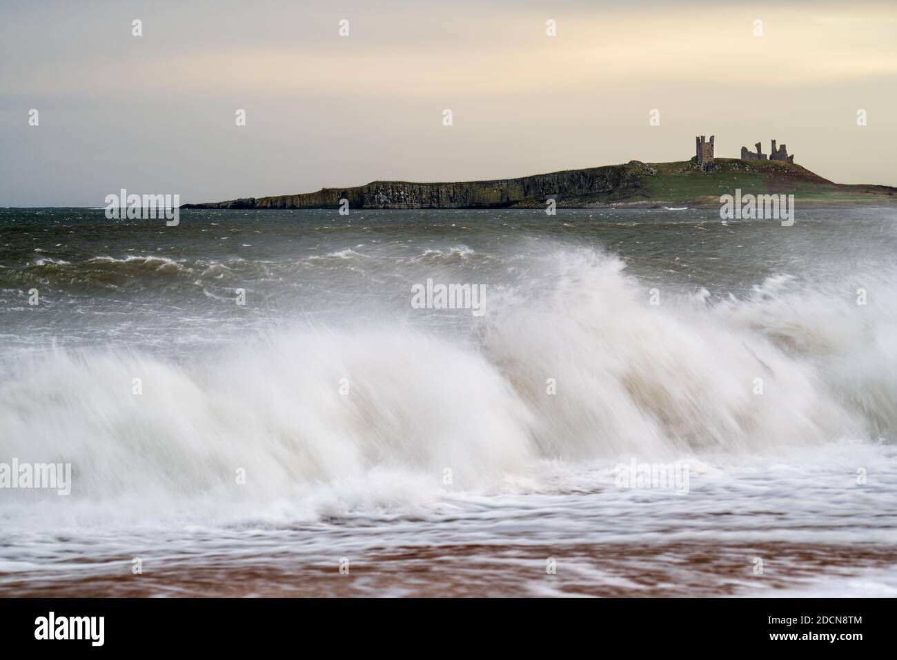 Dunstanburgh Castle liegt an einem windigen Nachmittag auf den Klippen über der Embleton Bay, wo große Wellen entlang der Küste brechen, wenn die Flut anrückt. Stockfoto