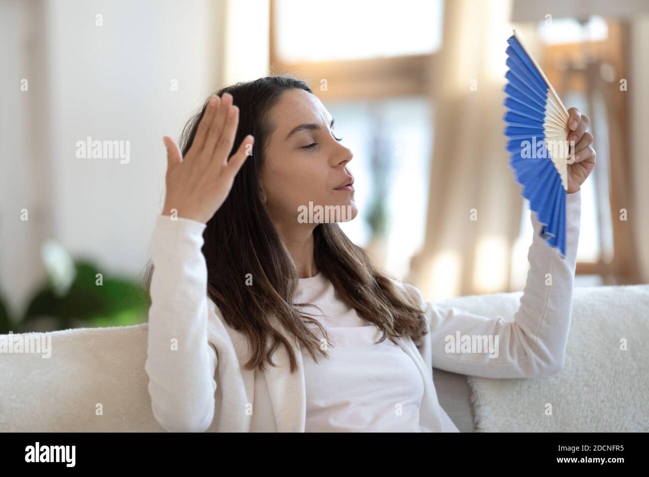 Gestresste Frau sitzt auf der Couch winkt sich mit Handventilator Stockfoto