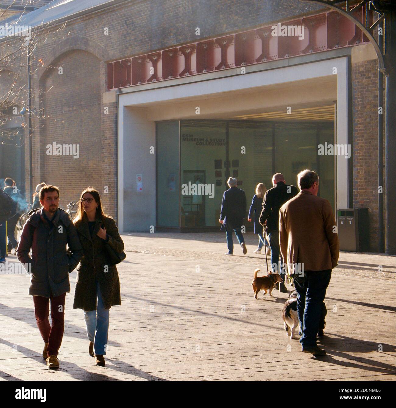 Junges Paar, das Arm in Arm zu einem Lebensmittelmarkt im Winter mit Leuten geht, die Hunde weg vom Markt gehen. Coal Drops Yard, Kings Cross, London. Stockfoto