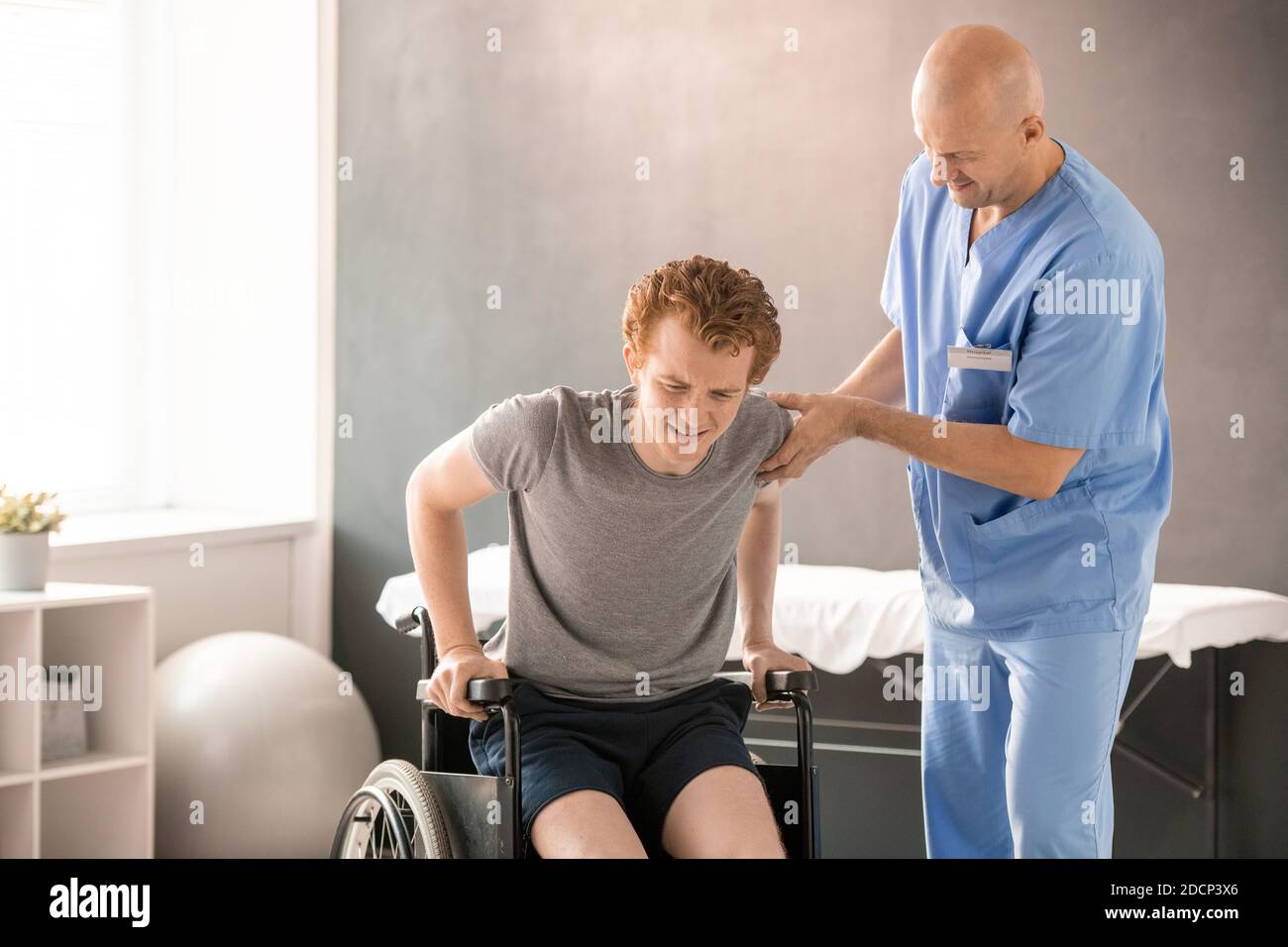 Reifer Arzt in Uniform hilft jungen Mann in Schmerzen zu Sitzen Sie im Rollstuhl Stockfoto